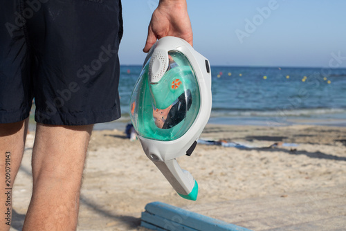 Man holding a full-face snorkel mask while walking along a sandy beach with clear blue water and distant swimmers in the background
