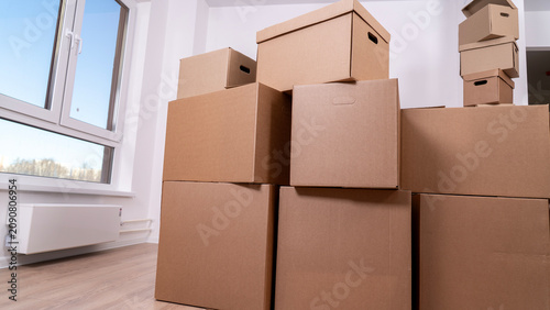 Stacked cardboard boxes occupy a bright room. Large window lets in natural light. Wooden floor shows clean space. Unopened boxes suggest packing or moving. Ideal for moving companies or storage themes