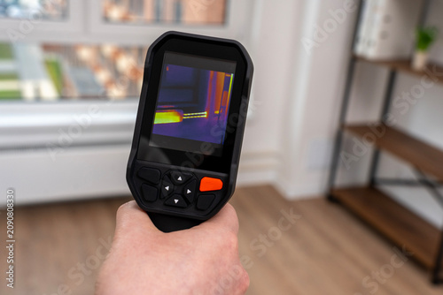 Hand holds black thermal camera. Screen shows color-coded heat pattern. Room features wooden floors and bookshelves. Window reveals blurred outdoor view. Ideal for home energy audits
