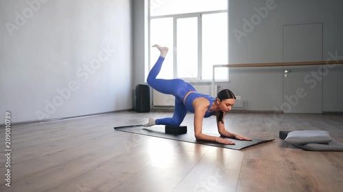 Graceful motivated fit attractive woman in activewear practicing fire hydrant exercise paired with donkey kick using yoga blocks, strengthening glute and hip muscles while training in fitness club.
