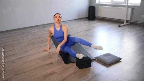 Graceful sporty fit woman practicing stretching exercises, doing front splits paired with seated forward bend using yoga blocks, improving flexibility and strengthening muscles while training in gym.