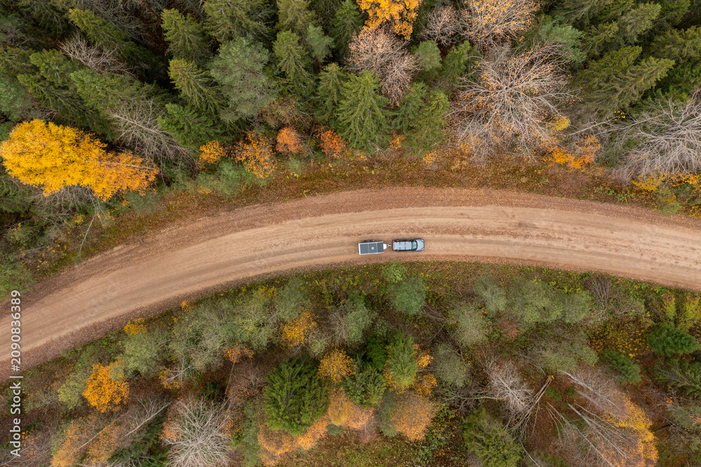 Naklejka premium Overhead drone view of an SUV with a camping trailer driving on a curving dirt road surrounded by dense autumn woods.