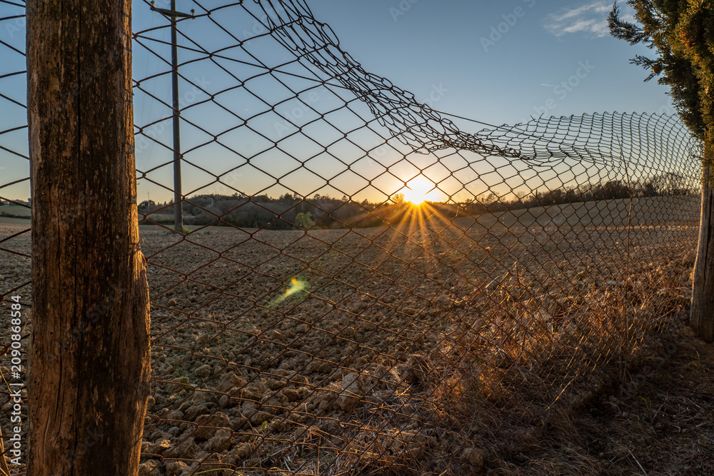 Obraz premium Sunset view over farmland seen through a netted field and wooden fence. The image captures the warm light and open rural atmosphere of Tuscany.