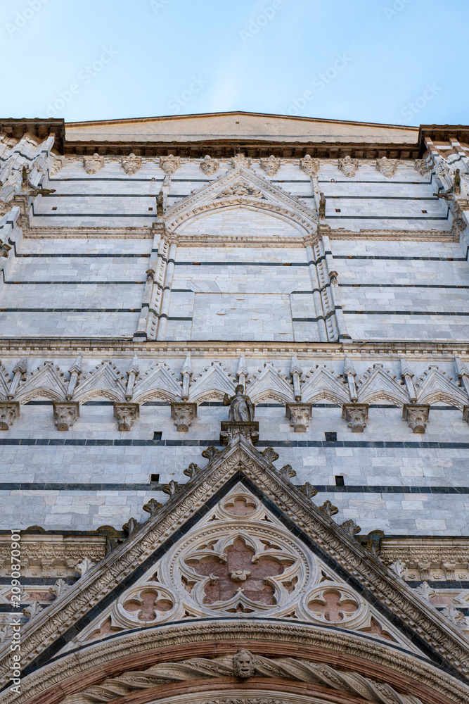 Fototapeta premium Close-up view of the ornate white marble facade of Siena Cathedral in Tuscany, Italy. The image highlights the intricate Romanesque and Gothic architectural details against a clear blue sky.