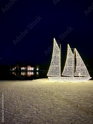 Illuminated Christmas decorations shaped like sails light up a snowy landscape at night in Trakai, Lithuania. In the background, the historic Trakai Island Castle