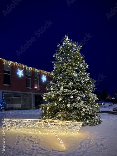 A large Christmas tree stands in Trakai, Lithuania, adorned with white ornaments and illuminated lights. It is surrounded by a snowy landscape. Nearby, a boat-shaped light decoration 