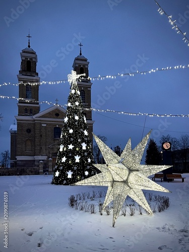 Large Christmas tree decorated with white star ornaments and lights stands in Vievis, Lithuania. In the foreground, an illuminated star sculpture is encircled by snow. Two church towers with crosses 