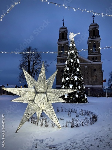 A decorated Christmas tree stands in a snowy square in Vievis, Lithuania, adorned with white star-shaped ornaments and illuminated by lights. In the foreground, a large, illuminated star sculpture 