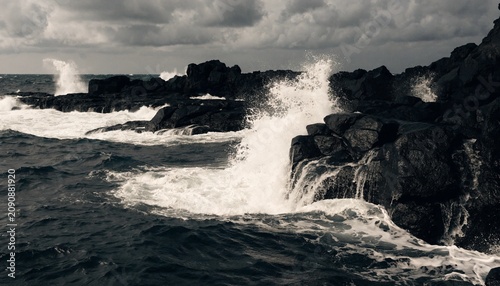 Dramatic rugged coastline with storm waves crashing over jagged black volcanic rocks, explosive white sea spray, dark ocean water, and moody overcast clouds in wild natural seascape