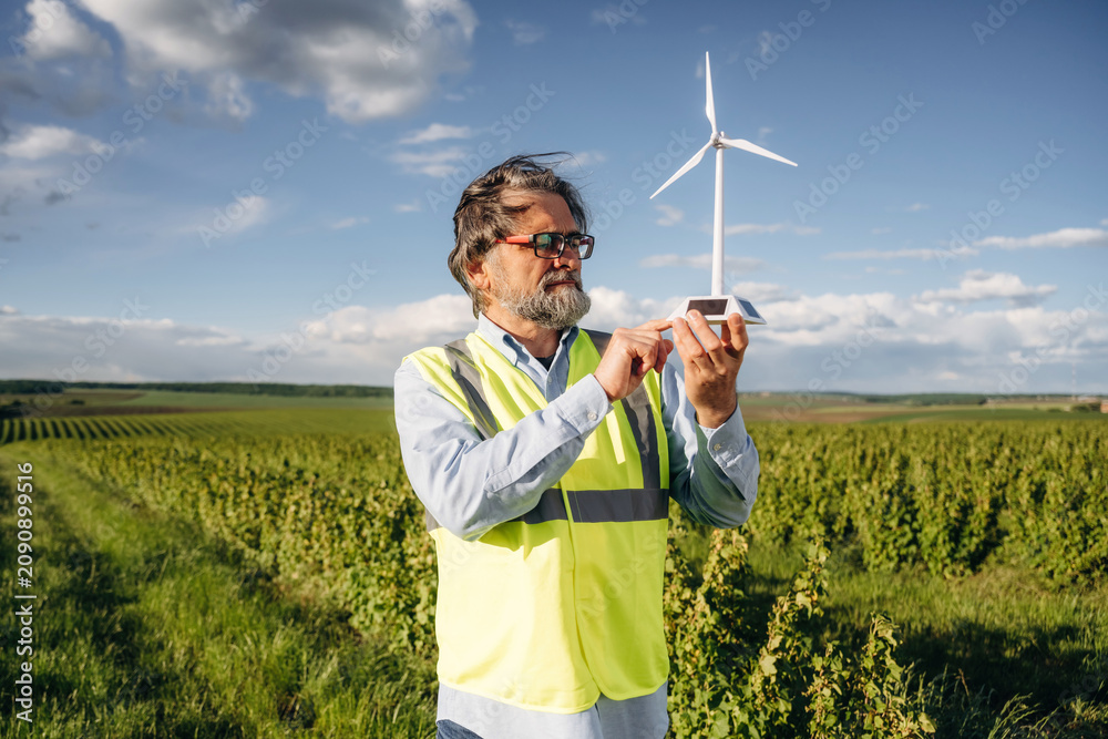 Fototapeta premium Middle-aged man in reflective vest examines small wind turbine model in green vineyard field under a blue sky with scattered clouds during daylight