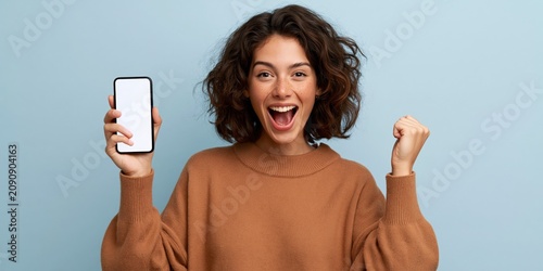 Excited curly haired woman holding a smartphone with a blank white screen on blue backdrop for app promotion and web banner