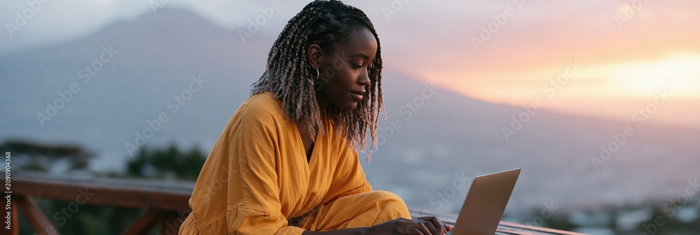 Fototapeta premium Young woman using laptop outdoors at sunset on balcony with mountain and city view for remote work background and banner design