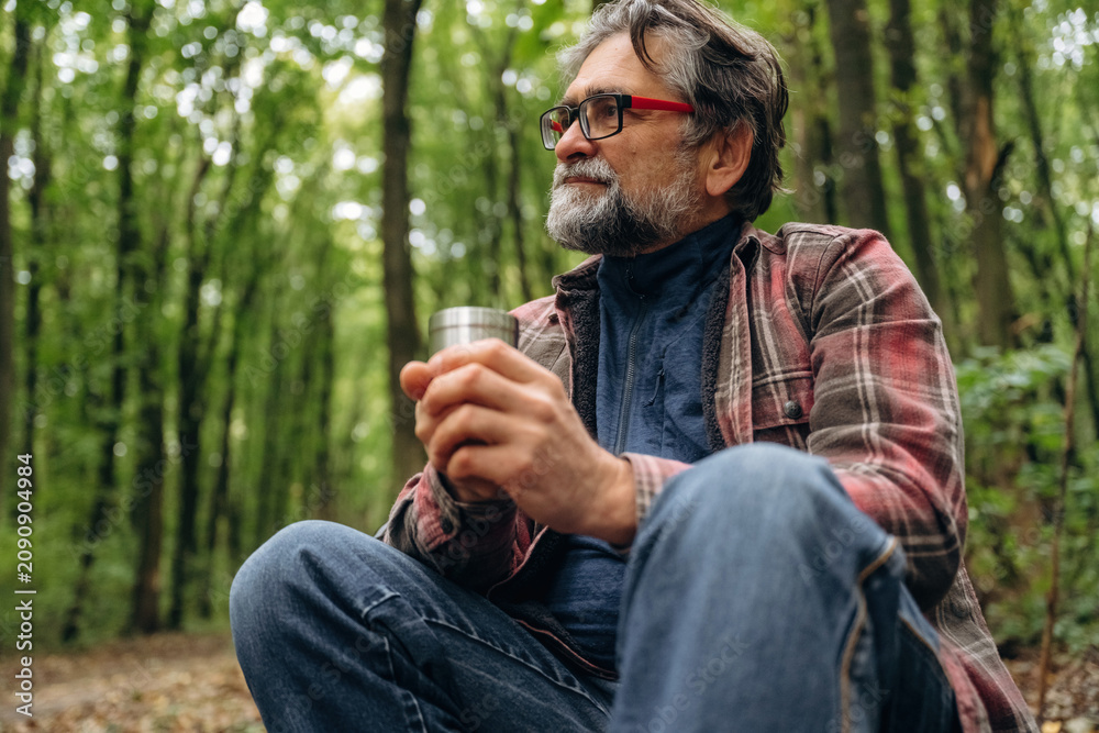 Fototapeta premium Enjoying the hot drink from thermos. Hiking experience, senior man is in the forest at daytime