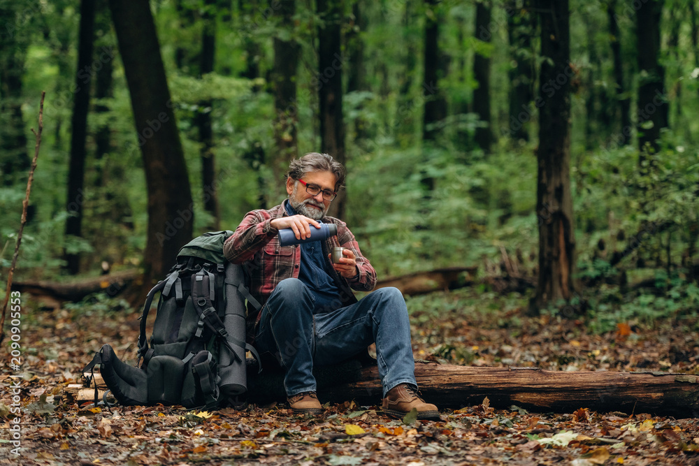 Fototapeta premium Enjoying the hot drink from thermos. Hiking experience, senior man is in the forest at daytime