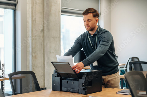 Fotografie Male office worker in gray sweater placing documents into a multifunction printe