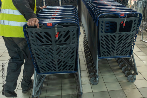 Supermarket employee in high-visibility safety vest collecting empty plastic shopping carts in retail store mall corridor, retail logistics and supermarket maintenance concept