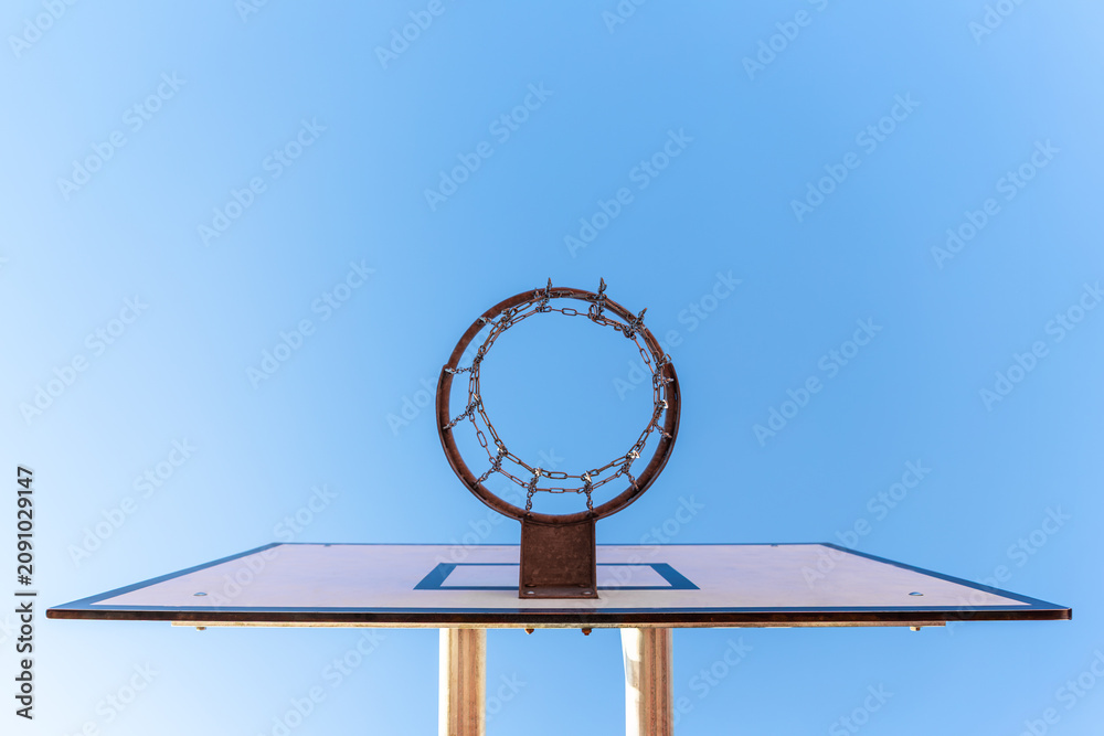 Fototapeta premium Outdoor basketball hoop with weathered metal rim, chain net, and backboard stands against clear blue sky. Low viewpoint creates urban street mood, minimal composition, sport energy, and summer play