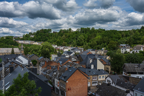 Panoramic aerial view of Houffalize town in Belgium, featuring traditional slate roofs, European architecture, and lush green hills under a cloudy sky.