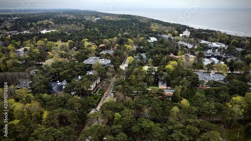 Aerial view of Palanga, Lithuania, showcasing a lush, densely forested area near the Baltic Sea coastline. Residential buildings are nestled among the trees, with varying architecture