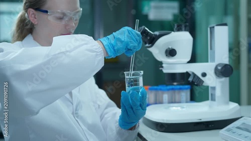 Female scientist mixing chemical liquid in beaker for experiment in scientific laboratory with microscope and test tubes