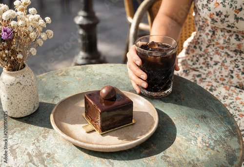 Elegant chocolate mousse cake with glossy glaze served on ceramic plate with iced black coffee in a glass. Outdoor cafe scene with female hand and soft natural sunlight.