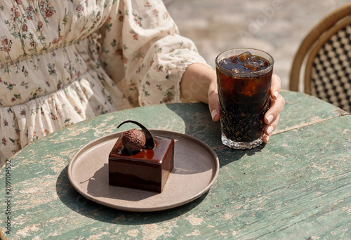 Elegant chocolate mousse cake with glossy glaze served on ceramic plate with iced black coffee in a glass. Outdoor cafe scene with female hand and soft natural sunlight.