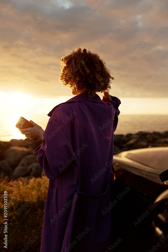 Fototapeta premium Young woman with curly hair stands beside a car at sunset, looking at her phone with a calm, reflective mood, conveying a casual lifestyle moment outdoors as warm light washes the scene and colors