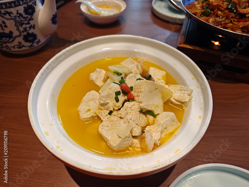 A white ceramic bowl filled with soft tofu cubes simmering in a savory golden broth, garnished with scallions and goji berries