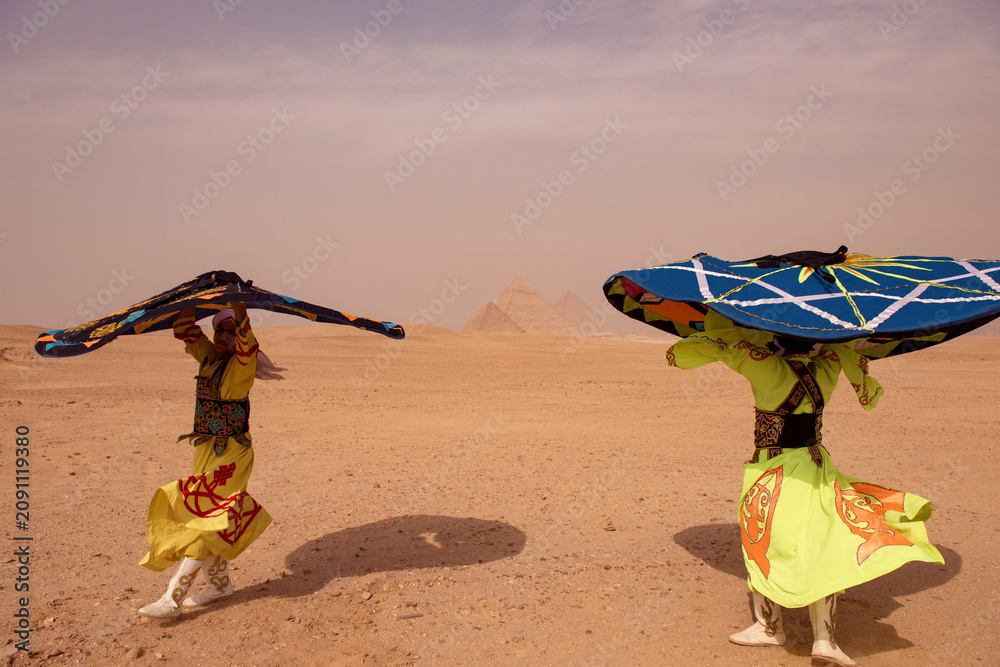 Fototapeta premium Dual tanoura performance dancers in neon yellow robes spinning enormous painted discs on Giza desert plateau with iconic pyramids centered between