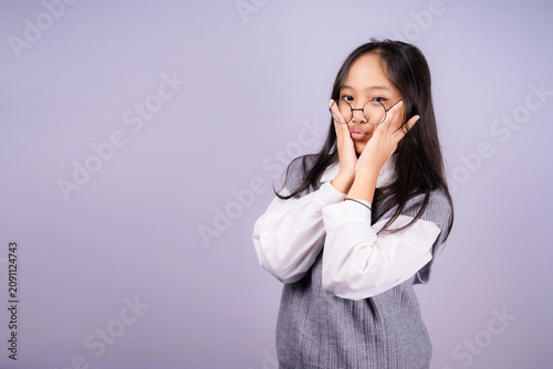 Schilderij op canvas Funny Asian schoolgirl in uniform with round glasses and puckered lips, pressing cheeks with hands, showing playful expression on light background, ready for back to school