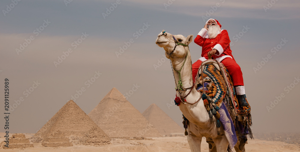 Fototapeta premium Camel rider dressed as Santa Claus throws head back in joyful belly laugh atop decorated dromedary with three ancient stone pyramids visible across