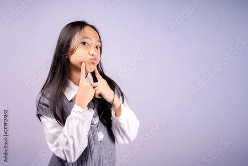 Schilderij op canvas Playful Asian schoolgirl in uniform making a funny face with puckered lips and fingers on cheeks, posing against light background, ready for back to school season