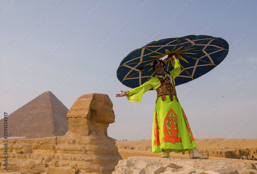 Fototapeta premium Sphinx performer in vivid neon robe raises large teal decorative disc overhead beside Great Sphinx at Giza plateau desert Egypt cultural tourism