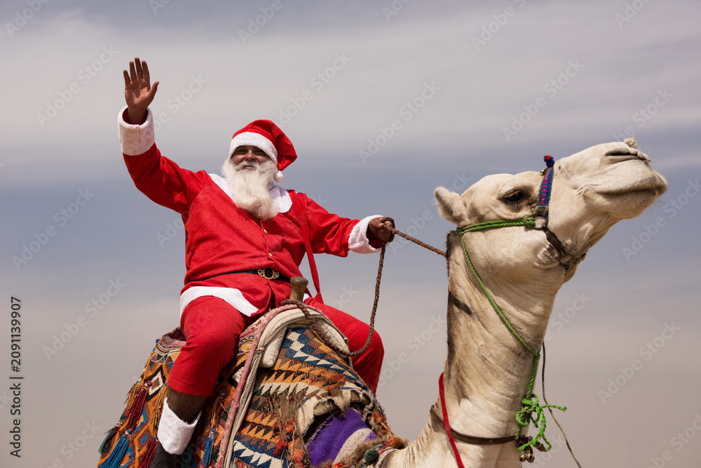 Fototapeta premium Santa waving enthusiastically from atop a white camel with vivid woven saddle blanket under pale desert sky ideal for global Christmas and