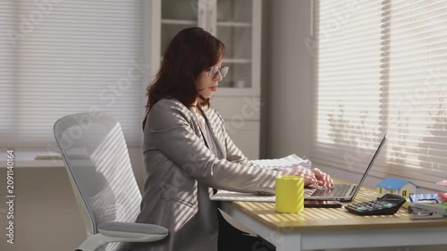 Businesswoman working on laptop at sunny office desk drinking coffee while typing on keyboard
