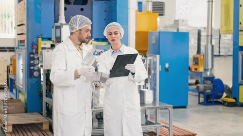 Male and female lab workers wearing protective coats and hairnets observing aluminum trays. Quality control specialists checking production process at modern industrial factory in tablet.