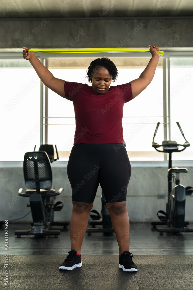 Fototapeta premium Plus size African American woman lifting green resistance band overhead in modern gym, fitness lifestyle photo for shoulder mobility, healthy workout routine, body confidence and wellness motivation.