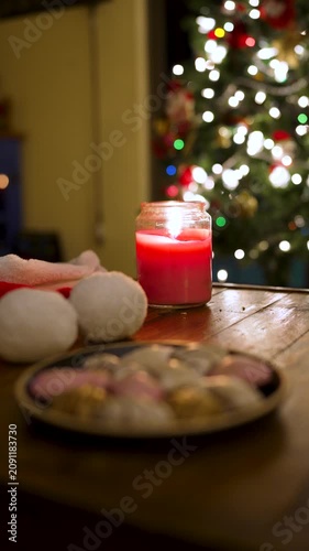 Red burning candle on a wooden table with cookies and a decorated christmas tree in the background