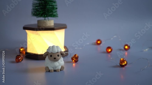 Vintage 2027 symbol white sheep figurine stands against the background of a glowing paper and wood lamp with a miniature Christmas tree on a background of flashing acorn-shaped garlands on a blue back