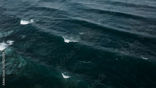 Aerial view of dark blue ocean waves crashing with white foam and ripples under a cloudy sky