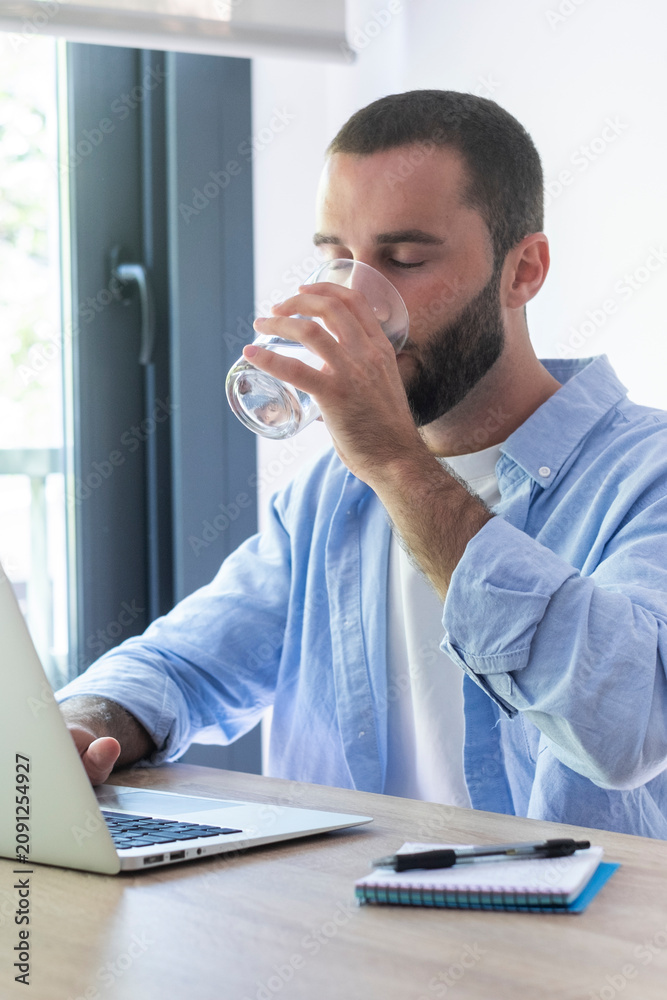 Fototapeta premium Man drinking water while working on laptop at home office, remote work, healthy routine and focused young professional