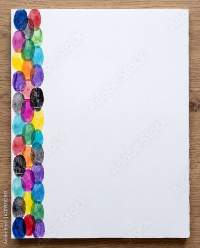 Vertical border of colorful fingerprint stamps on white watercolor paper, shot on a wooden desk with ample copy space. Ideal for posters, invitations, stationery, branding templates, and web design.