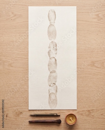Minimal vertical column of neutral fingerprints on white paper, arranged on a wooden desk with pen, brush and wax cup. Clean forensic or security background with copyspace for design.