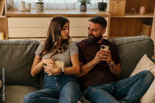 Фотография Woman feeling jealous observing man using phone on sofa