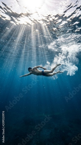 A serene underwater perspective of a professional swimmer gliding through the deep blue ocean, illuminated by ethereal sunbeams piercing the surface in a display of athletic grace and aquatic harmony.