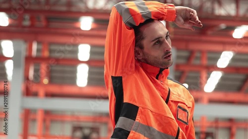 Exhausted industrial worker in high-visibility orange jacket wiping sweat from his forehead. Warehouse worker showing signs of physical fatigue and heat strain.