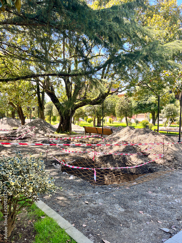 Construction site in sunny park under large trees with taped-off pit.