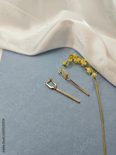 Modern gold bar drop earrings on light blue background. Elegant jewelry flat lay with white satin and dried plant