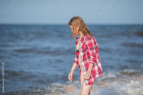 Girl in red plaid shirt standing by sea waves on beach.