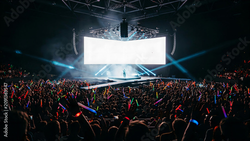 Concert crowd holding glowing light sticks in a large dark arena with a bright stage and spotlights